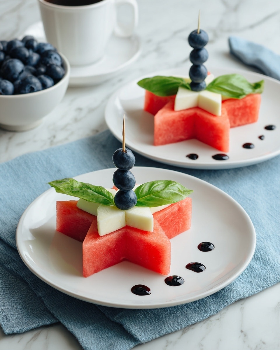 The dish shows a white plate on a white marbled surface with a star-shaped slice of red watermelon as the base layer. On top of the watermelon, there is a smaller white star-shaped piece, likely cheese, centered in the middle. A small dark blueberry is placed on top of the white piece, held in place by a toothpick. Bright green basil leaves are positioned between the white piece and the blueberry. To the side of the star shapes, five small dots of dark sauce are lined up neatly on the plate. Another similar plate is visible in the background, and a white cup filled with blueberries sits nearby. The scene is set on a blue fabric cloth. Photo taken with an iphone --ar 4:5 --v 7