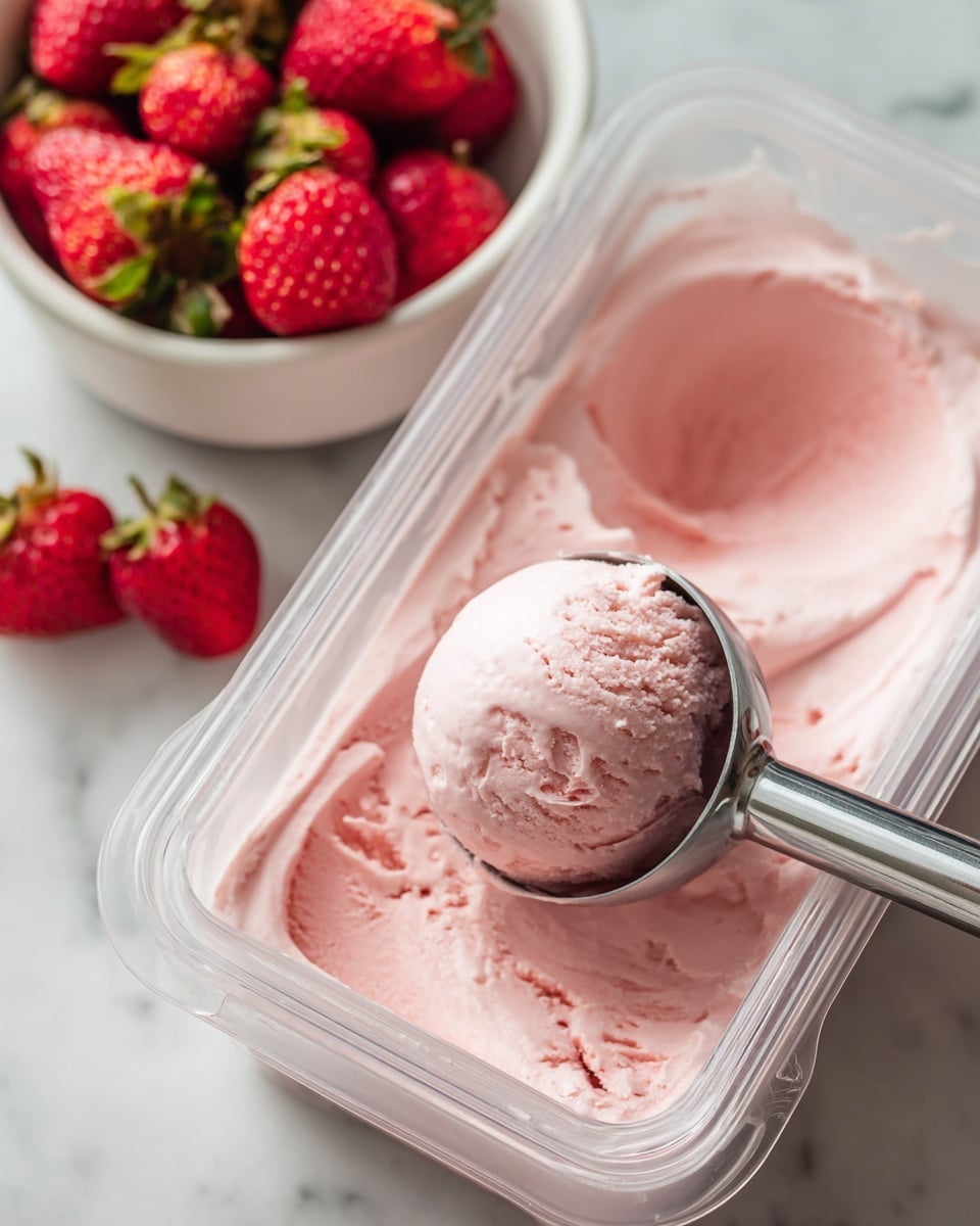 A clear ice cream scoop is holding a smooth, thick scoop of light pink creamy ice cream inside a transparent ice cream maker container showing the creamy texture clearly. The scoop sits in the middle of the container filled with gently swirled ice cream, while next to it there is a white bowl containing bright red fresh strawberries with green tops. The background is a white marbled surface with soft light bringing out the bright colors and smooth textures. photo taken with an iphone --ar 4:5 --v 7