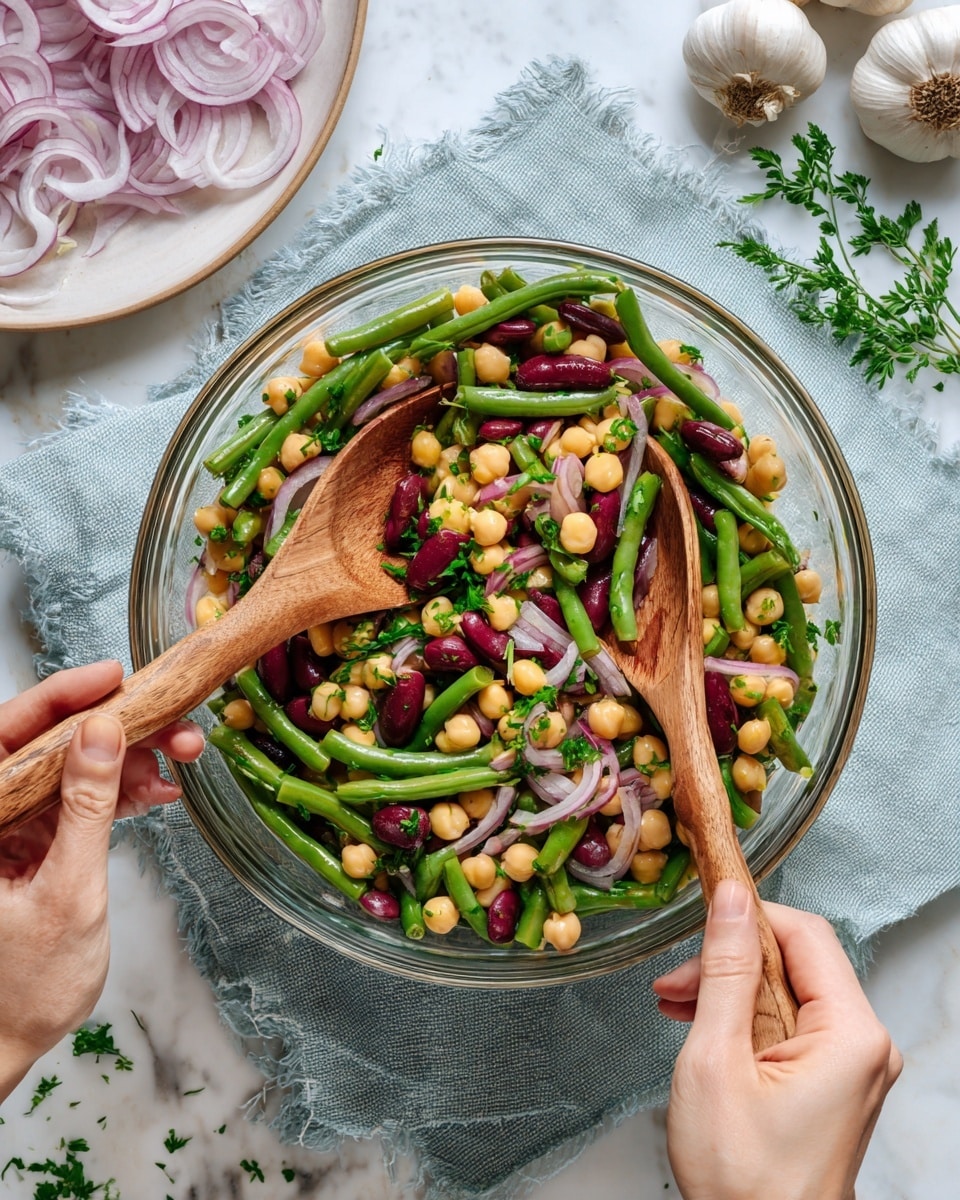 A clear glass bowl filled with a colorful salad sits on a white marbled surface. The salad has three main layers: a base of green beans cut into small pieces, a middle layer of dark red kidney beans, and light beige chickpeas mixed throughout. Thin slices of purple onion curls rest on top, adding a light contrast. The salad is sprinkled with small green herb pieces, giving it a fresh look. Two wooden spoons stir the salad, held by a woman's hand on each side. Nearby, there is a white plate with more onion slices, a garlic bulb, a small glass bottle of olive oil, and fresh green parsley leaves. Photo taken with an iphone --ar 4:5 --v 7