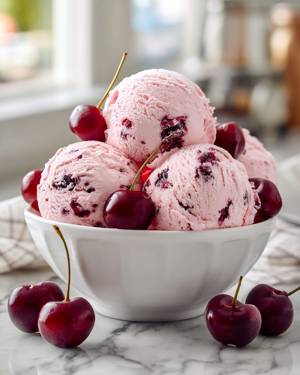 A white bowl filled with four round scoops of light pink cherry ice cream with visible dark cherry pieces inside. On top and around the ice cream scoops are several whole dark red cherries with shiny skin, some touching the bowl's edge. The bowl is placed on a white marbled surface with three loose cherries beside it. The background is softly blurred, showing a kitchen with light coming through a window. Photo taken with an iphone --ar 4:5 --v 7