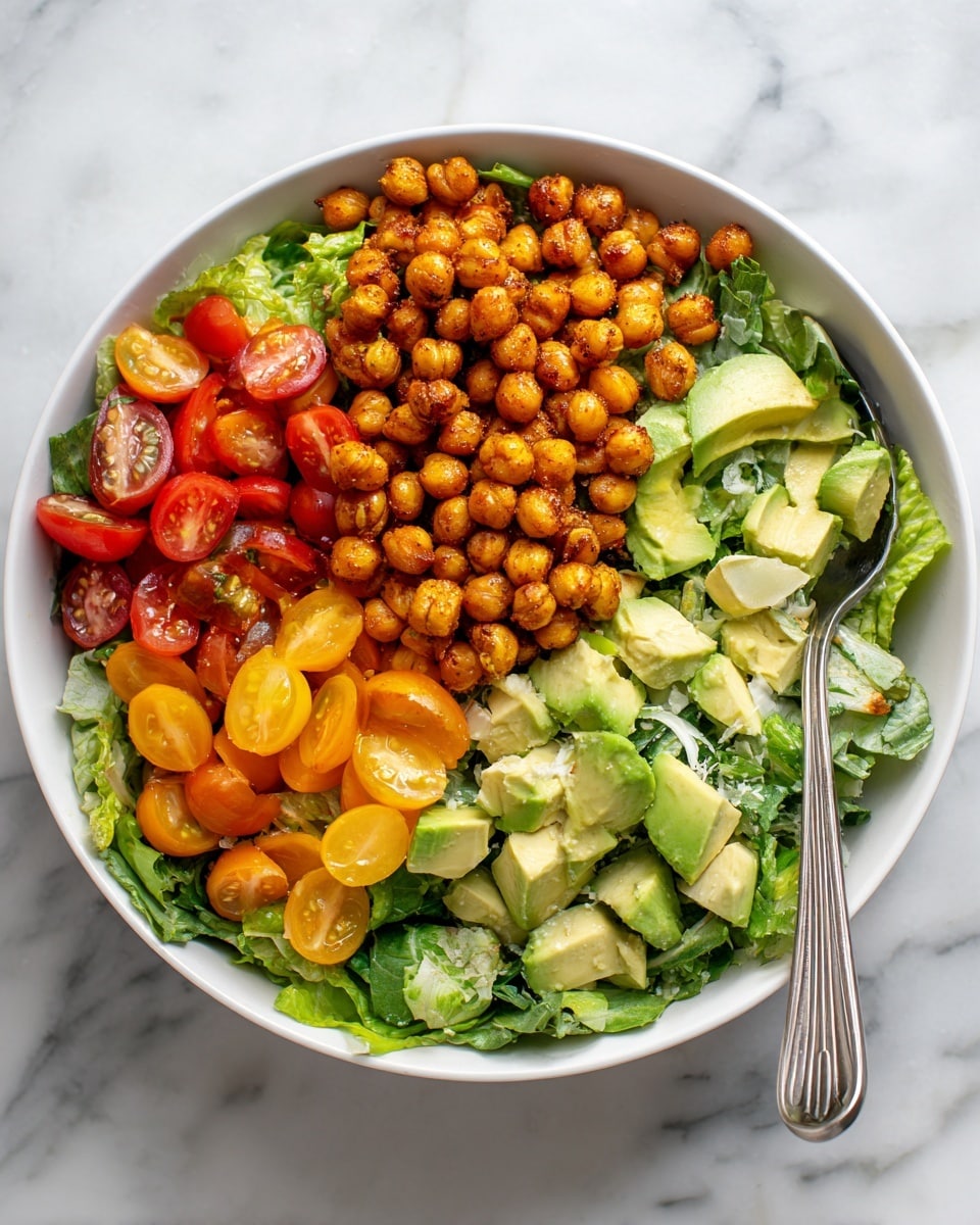 A white bowl holds a colorful salad with four main layers: the bottom layer is fresh green and purple leafy lettuce, topped with chopped red and yellow cherry tomatoes and light green avocado chunks scattered evenly; the third layer is sliced cucumber pieces mixed throughout, and the top layer is a generous amount of golden-brown roasted chickpeas covering the salad; some white seeds are sprinkled on top. A metal spoon stands inside the bowl on the right side. The bowl sits on a white marbled surface. Photo taken with an iphone --ar 4:5 --v 7