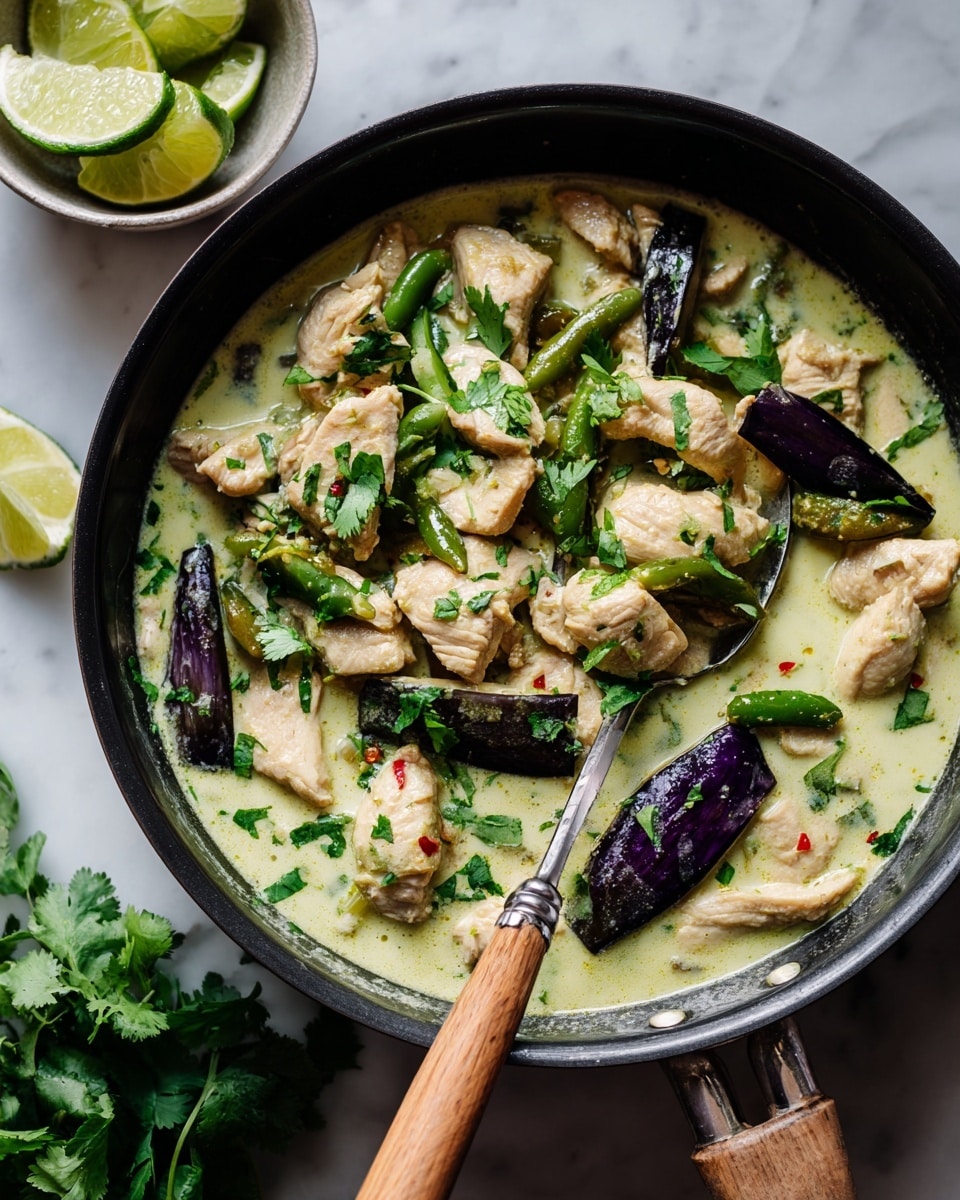 The image shows a black skillet filled with a creamy light green curry with layers including chunks of light beige chicken, slices of dark purple eggplant with white insides, and long green chili peppers, all floating in a thick curry sauce. On top, there are bright green coriander leaves scattered for garnish. A spoon with a wooden handle is scooping some curry with chicken and eggplant. Next to the skillet, a white bowl holds wedges of fresh lime. The surface under the skillet is a white marbled texture. Photo taken with an iphone --ar 4:5 --v 7