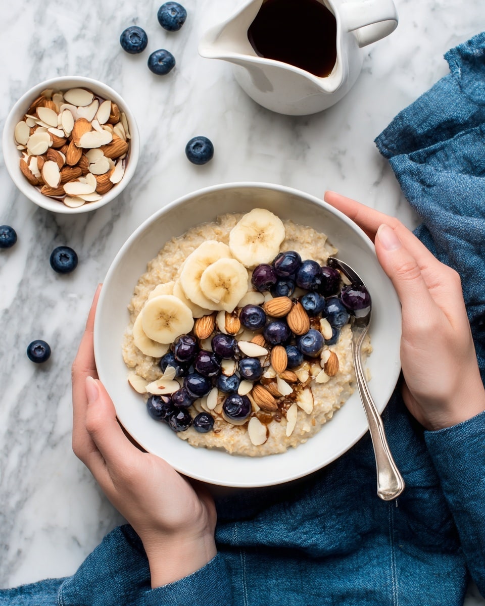 A beige bowl filled with a smooth oatmeal base forms the bottom layer. On top, there are round banana slices with a pale yellow color, dark blue blueberries, and chopped brown almonds scattered evenly. A woman's left hand is holding the bowl from the side while her right hand holds a spoon scooping into the oatmeal. The bowl sits on a white marbled surface with a white small bowl of chopped almonds and a white small pitcher with dark syrup in the background. Dark blue blueberries are scattered around the bowl and there is a blue-striped cloth underneath one side. Photo taken with an iphone --ar 4:5 --v 7