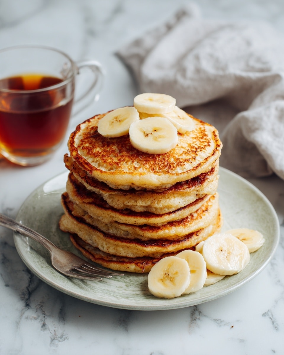 A stack of six round pancakes with a golden brown and slightly crispy texture is centered on a white plate. Each pancake has a warm, uneven browning, with the middle pancakes darker in shade. On top of the stack, there are two slices of pale yellow banana, and to the side of the plate, more banana slices are arranged. A silver fork rests on the left side of the plate. In the background, a clear glass cup filled with amber-colored syrup sits on a white marbled surface, next to a soft gray cloth. Photo taken with an iphone --ar 4:5 --v 7