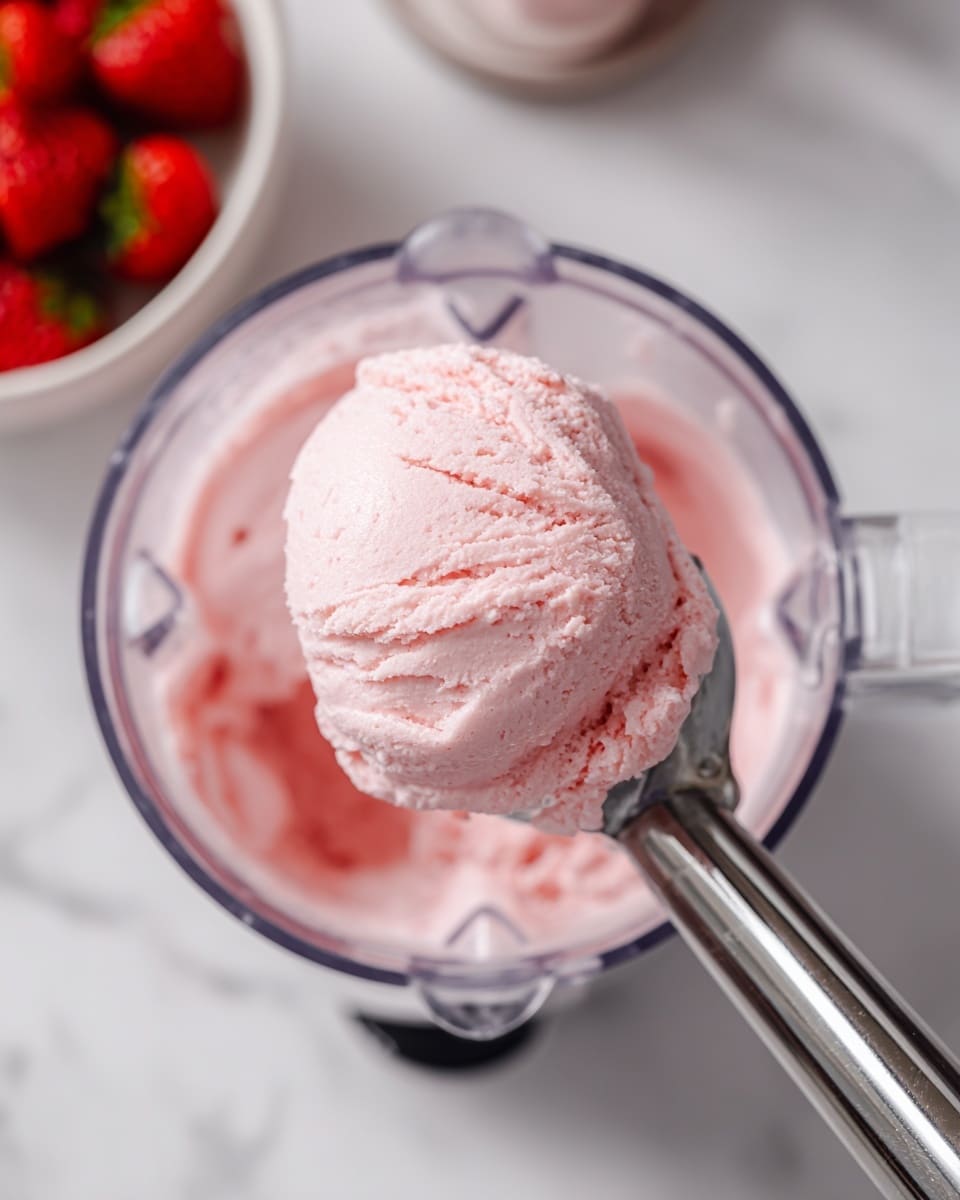 A close-up image showing a clear blender container filled with smooth, creamy light pink ice cream. A metal ice cream scoop is inside the blender, lifting a rounded, soft scoop of the pink ice cream. In the background, a white bowl filled with bright red strawberries is partly visible, sitting on a white marbled surface. The top edge of the blender lid is seen to the left side of the image. photo taken with an iphone --ar 4:5 --v 7