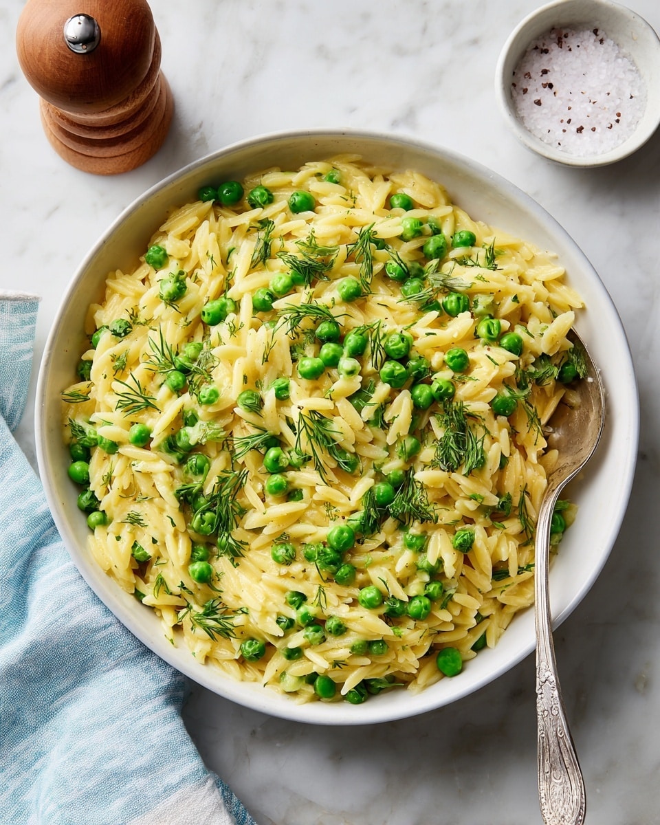 A white bowl filled with creamy yellow orzo pasta mixed with bright green peas, giving a soft and slightly shiny texture. The pasta is evenly coated with a smooth sauce and topped with fresh green herbs like parsley and dill, adding a leafy texture and color contrast. A silver spoon rests inside the bowl, ready for serving. The bowl sits on a white marbled surface, with a small white bowl of coarse salt and a wooden pepper grinder nearby, along with a blue and white striped cloth partially visible. photo taken with an iphone --ar 4:5 --v 7