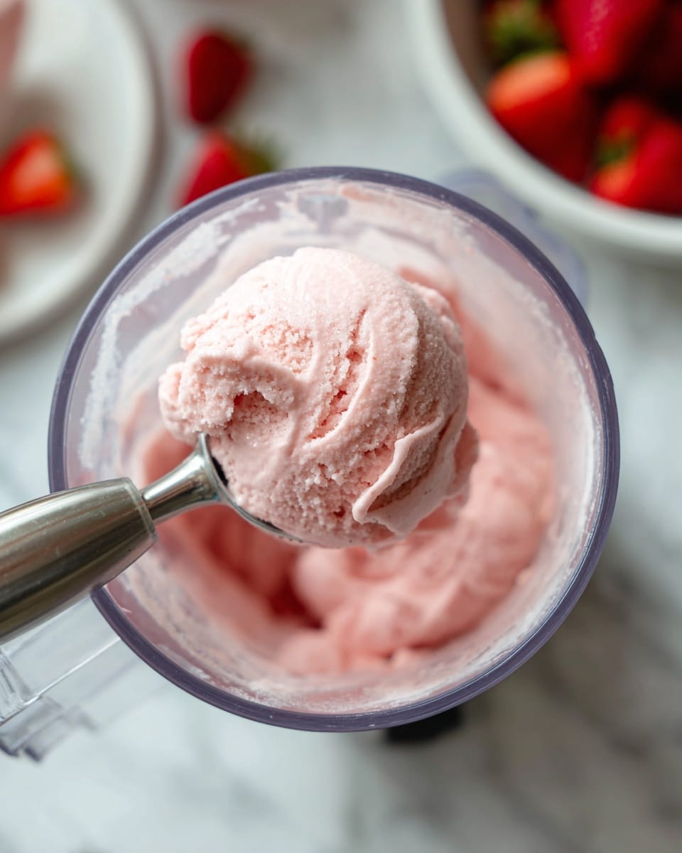 A close-up of a clear container inside a food processor filled with smooth, creamy light pink ice cream. A stainless steel ice cream scoop is lifting a round, soft scoop of the ice cream, showing its thick, smooth texture. In the background, there is a white bowl filled with fresh red strawberries sitting on a white marbled surface. The cold and creamy ice cream contrasts with the bright, fresh strawberries, and the whole scene is bright and clean. photo taken with an iphone --ar 4:5 --v 7
