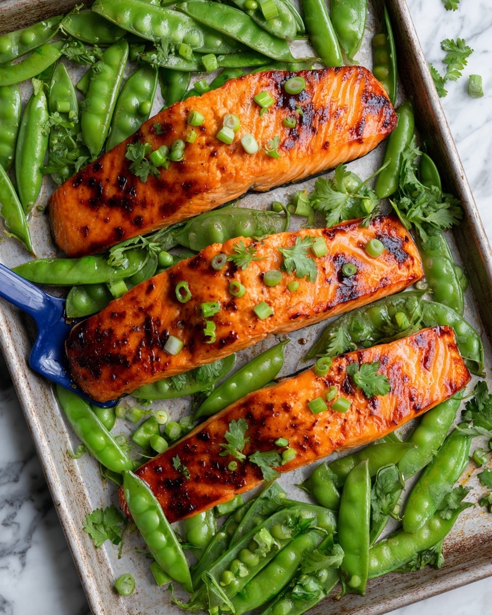 The image shows a metal baking sheet filled with three cooked salmon fillets that have a shiny orange-brown glaze on top with small darker grilled spots. Surrounding the salmon are many green snap peas that look slightly roasted and shiny with some small slices of cooked garlic mixed in. The salmon pieces are garnished with small green herb leaves and chopped green onions scattered over and around them. The background surface is a white marbled texture. photo taken with an iphone --ar 4:5 --v 7