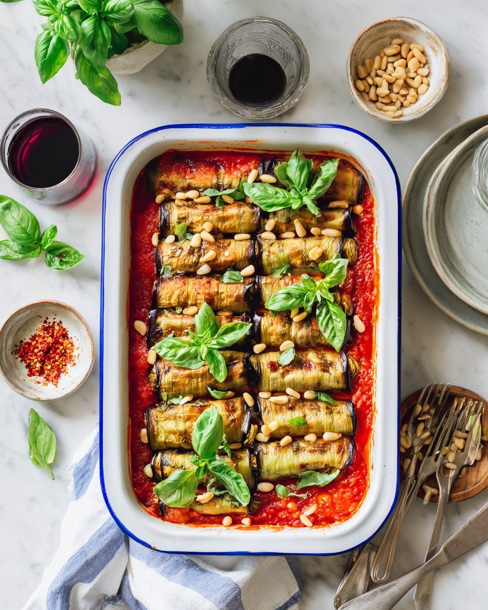 The image shows a white rectangular baking dish filled with layered eggplant slices that have a golden-brown grilled texture, arranged in a neat pattern on top of a rich red tomato sauce. The sauce layer underneath is visible between the eggplant pieces. There are fresh green basil leaves scattered on top along with light-colored pine nuts sprinkled evenly. The dish sits on a white marbled surface, next to a small gray plate with red chili flakes, a bunch of fresh basil, a beige bowl filled with pine nuts, and a clear glass of water. The scene is bright and clean with natural lighting, photo taken with an iphone --ar 4:5 --v 7
