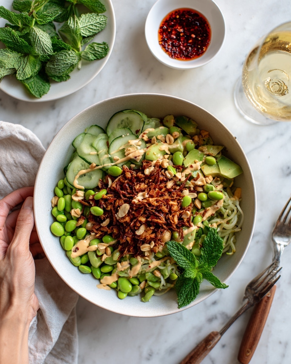 A white round bowl filled with a fresh salad featuring layers of sliced cucumber, chopped avocado, and green edamame beans spread evenly as the base, topped with crunchy golden-brown toasted grains and scattered light-colored peanuts. Light beige dressing is drizzled over the salad, with bright green fresh mint leaves placed on top and around the dish. The bowl sits on a white marbled surface, accompanied by a white plate holding more mint leaves and a small sauce dish filled with red chili flakes. Nearby there is a glass of a light yellow drink and a fork and knife with wooden handles. Photo taken with an iphone --ar 4:5 --v 7