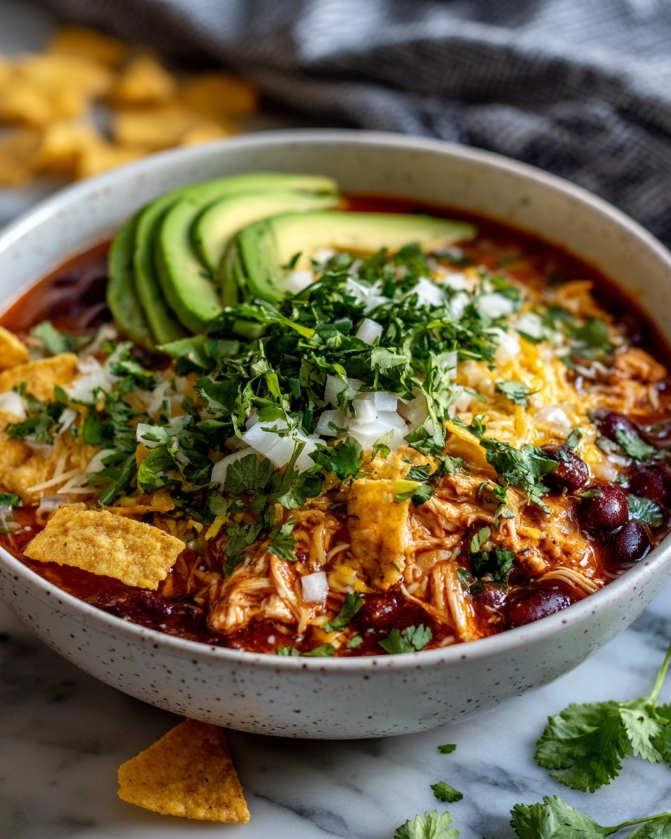 A white speckled bowl filled with a layered chili dish, showing shredded orange chicken mixed with dark red kidney beans in a red sauce base, topped with melted yellow cheddar cheese, small pieces of white onion, and fresh cilantro leaves. Two curved slices of green avocado rest on opposite edges of the bowl. The bowl sits on a white marbled surface with a gray and white striped cloth and yellow tortilla chip pieces scattered around. Photo taken with an iphone --ar 4:5 --v 7