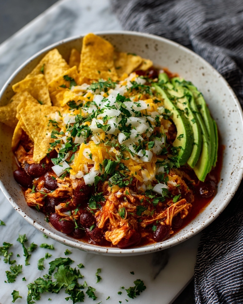 A round white bowl filled with a deep red chili soup base, layered with shredded orange chicken mixed with dark red kidney beans. This is topped with melted yellow and white cheese that blends smoothly into the soup. Bright green cilantro leaves and small cubes of white onion are scattered over the top, adding pops of color. Two fanned slices of light green avocado sit on the edge of the bowl. Some yellow tortilla chip crunch is sprinkled on the chili. The bowl rests on a white marbled surface next to a gray striped cloth with scattered broken yellow tortilla chips. photo taken with an iphone --ar 4:5 --v 7
