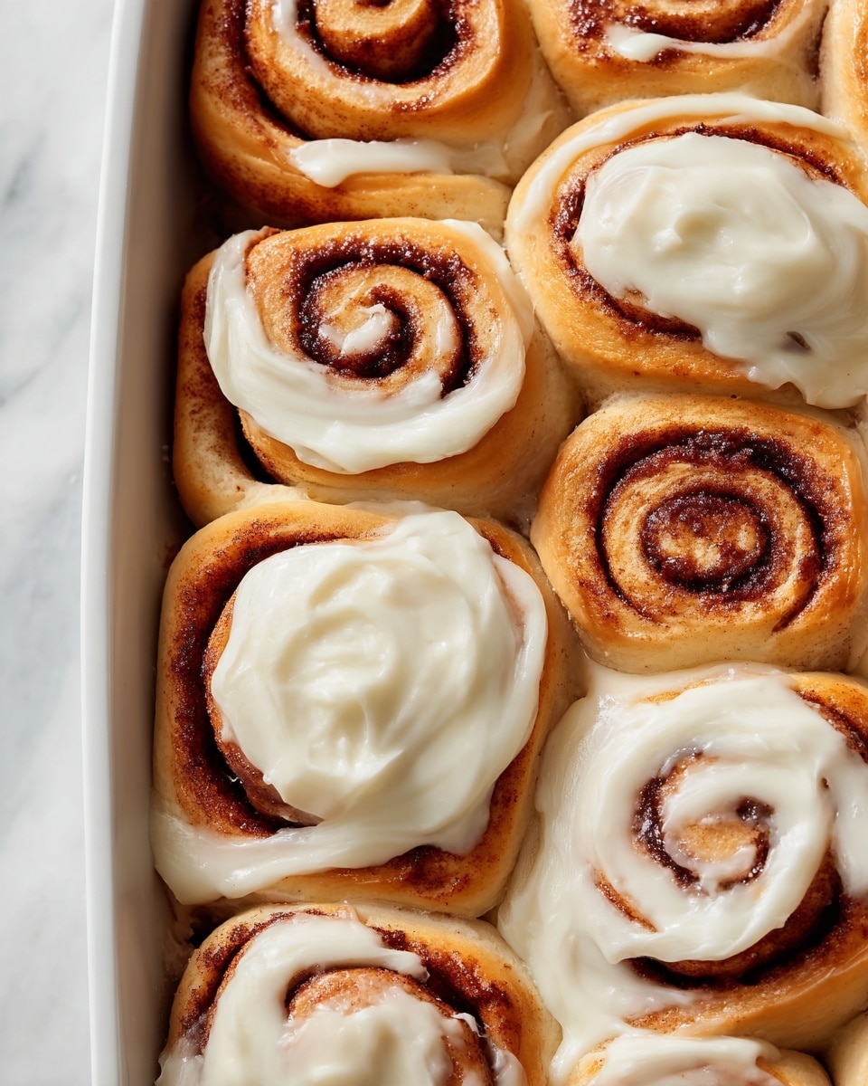 The image shows eight cinnamon rolls closely placed in a grid, with four of them covered in smooth, creamy white frosting spread unevenly, revealing the warm golden-brown rolls underneath. The rolls have swirled layers of dark brown cinnamon filling that contrasts against the soft, golden dough. The texture of the frosting is thick and slightly shiny, while the cinnamon layers look moist and sticky. The rolls fill the entire frame and sit on a white marbled surface. photo taken with an iphone --ar 4:5 --v 7
