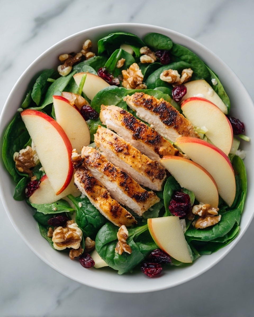 A white bowl filled with dark green leafy spinach at the base, layered with slices of grilled golden-brown chicken in the center. To the right, red apple slices are fanned out, and to the left, yellow pear slices are also fanned. Scattered around the greens are small white cheese chunks, bright red dried cranberries, and light brown walnut pieces. A silver fork rests at the top inside the bowl. The background is a white marbled surface with a glass container of yellow dressing on the right and a small bowl of walnuts at the bottom right. photo taken with an iphone --ar 4:5 --v 7
