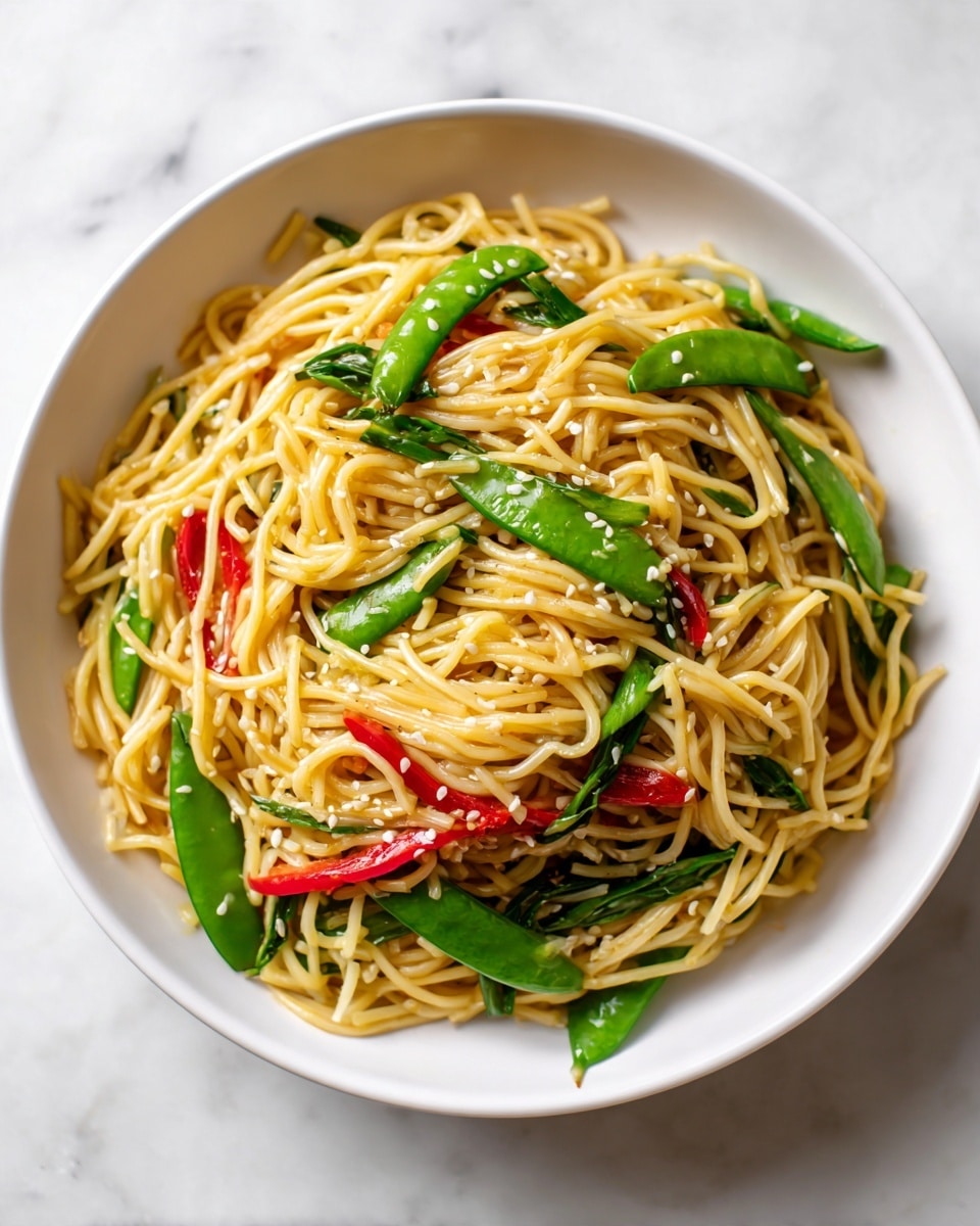 A white plate holds a colorful stir-fried noodle dish with thick light brown noodles as the base layer. Mixed in are bright green snap peas and long green onion pieces, scattered throughout the noodles. Bright red bell pepper strips add a pop of color on top. The whole dish is sprinkled with small white sesame seeds, adding texture and contrast. The background is a white marbled surface. photo taken with an iphone --ar 4:5 --v 7