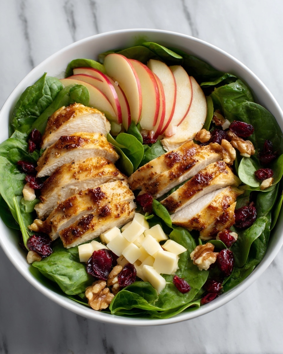 A white bowl filled with fresh green leafy spinach as the base layer, topped with two sections of thinly sliced grilled chicken with a light brown charred texture, positioned in the center. To the right of the chicken are thin slices of red apple with red skin still on, and to the left are thin slices of yellow pear with green skin. Scattered over the greens and fruit are small chunks of white goat cheese, whole walnut pieces, and dried red cranberries. A silver fork rests on top near the chicken and apple slices. The bowl is placed on a white marbled surface with a small glass container of yellow dressing and another bowl of walnuts nearby. Photo taken with an iphone --ar 4:5 --v 7
