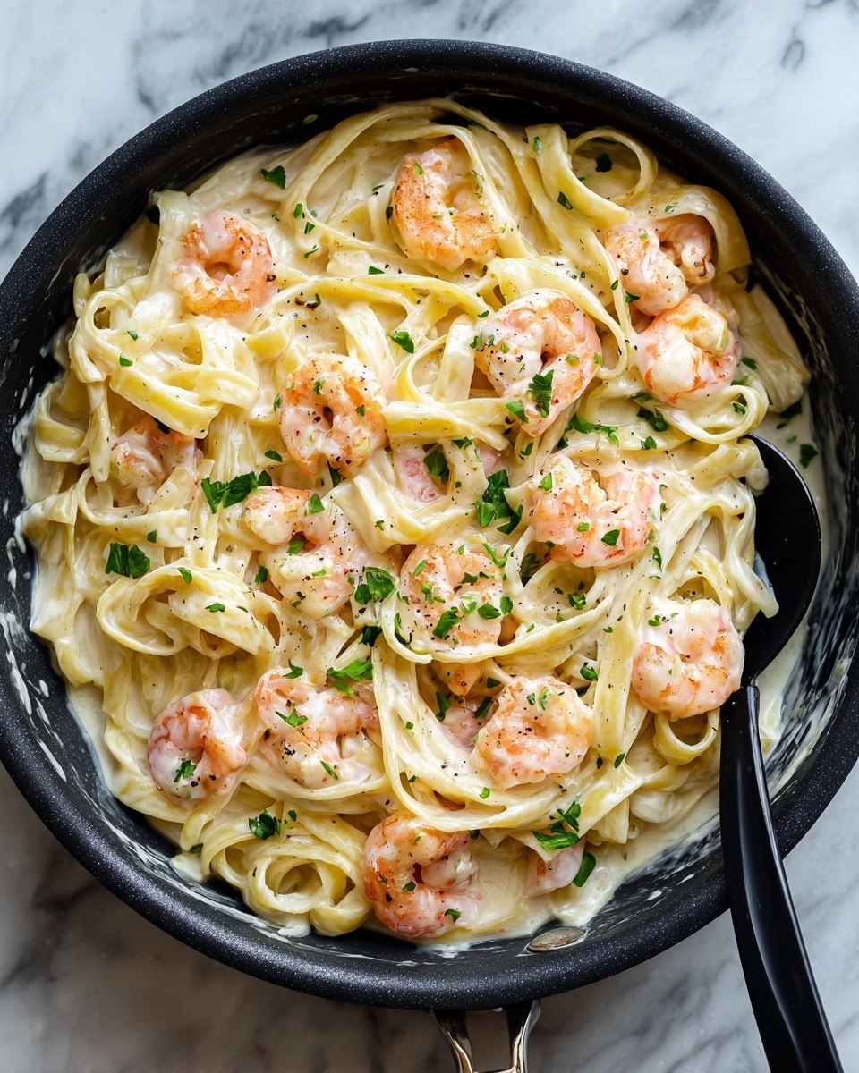 The image shows a black skillet filled with creamy white fettuccine pasta mixed evenly with light pink shrimp. The pasta strands are smooth and coated in a thick, white sauce that looks rich and silky. Small green herb pieces are scattered on top for color contrast. A black spoon rests on the right side of the skillet, partially submerged in the pasta. The skillet sits on a white marbled surface, highlighting the creamy colors of the dish. photo taken with an iphone --ar 4:5 --v 7