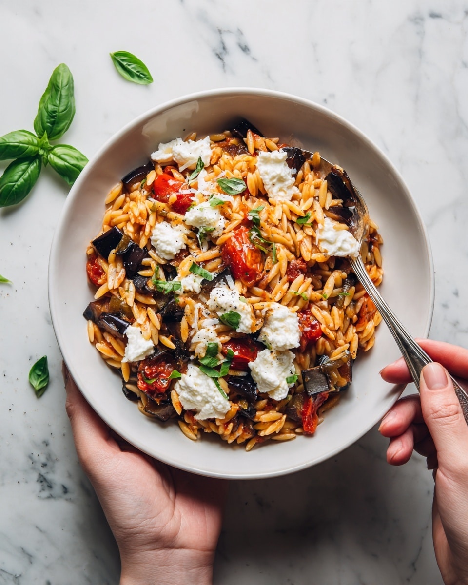 A bowl filled with reddish-orange orzo pasta mixed with small pieces of dark olives and bright green basil leaves, topped with white dollops of soft cheese. The pasta looks saucy and moist, with the cheese melting slightly on top. There are large fresh basil leaves scattered on the surface and a spoon inside the bowl. Two woman's hands are holding the bowl from opposite sides. The bowl is white, and the background has a white marbled texture. Photo taken with an iphone --ar 4:5 --v 7